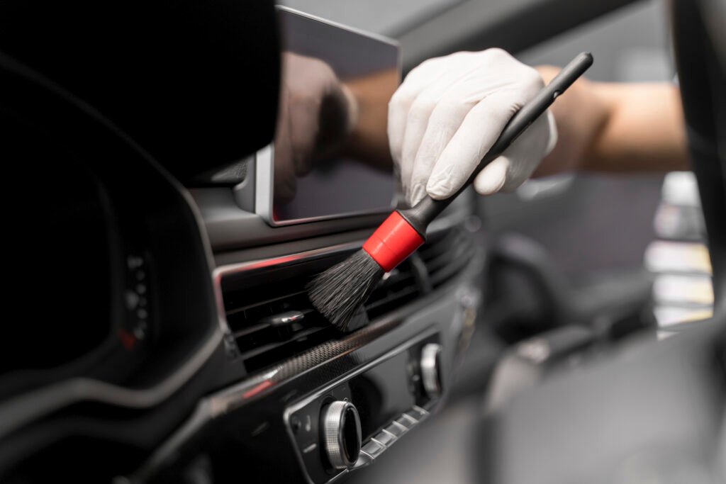 Professional car detailing service cleaning the interior dashboard and air vents using a specialized brush.
