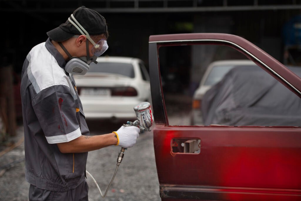 Image of a worker utilizing a professional air-powered spray gun for automobile paint restoration, demonstrating safety measures like a respirator.
