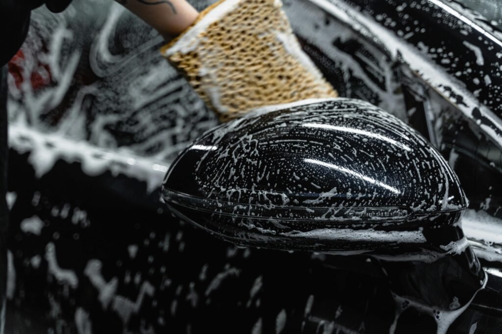 Image depicting a car wash service, where a worker is manually cleaning the exterior of a black car with a sponge and thick soap suds.